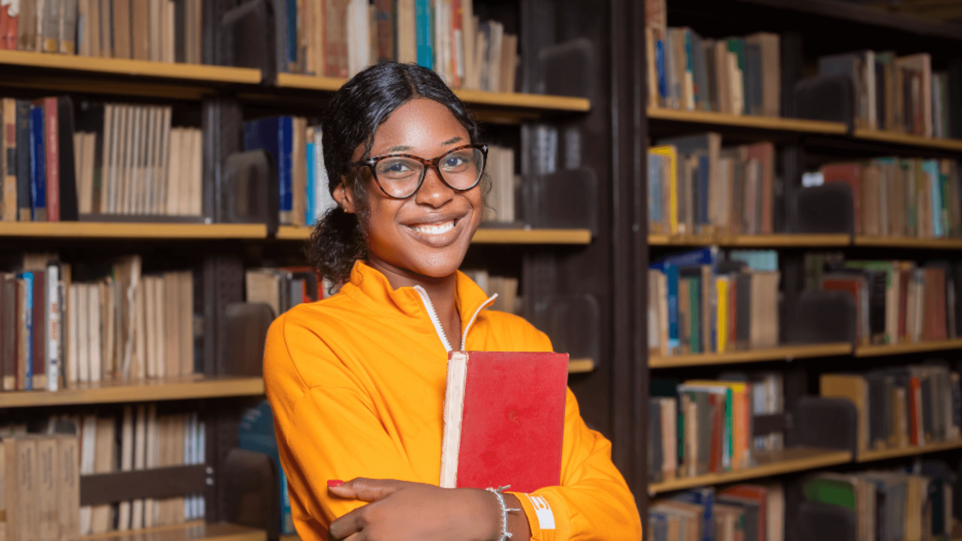 A young woman wearing glasses stands in front of a library, holding a book and looking thoughtfully at the camera.